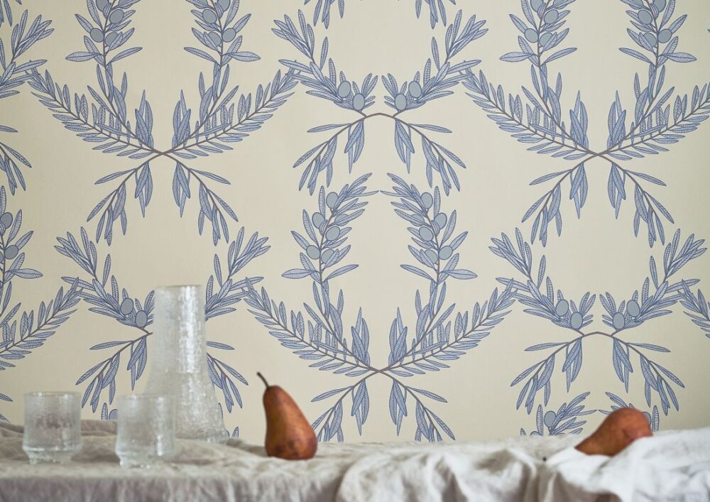Amalfi-Lines Oliva wallpaper with a soft blue olive branch design on a light beige background, paired with elegant glassware and a pear for a natural, sophisticated interior.