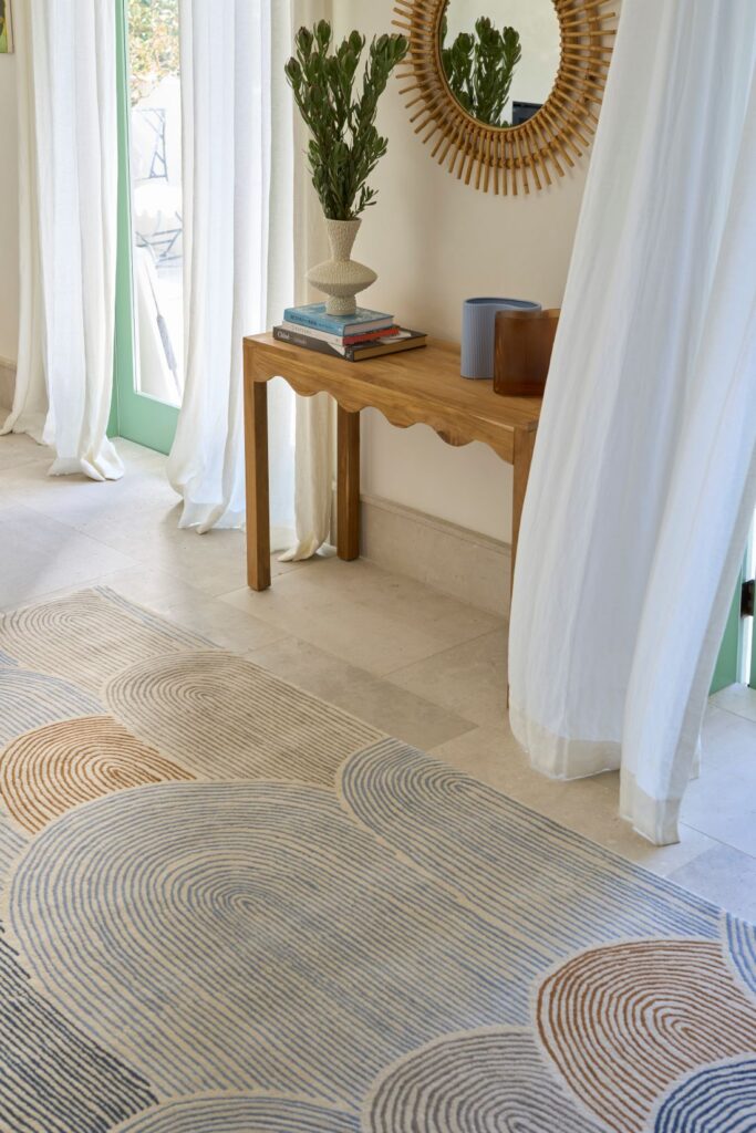 A stylish entryway featuring a wavy patterned rug in soft blues and terracotta tones, a wooden console table with decorative items, and a round mirror with a sunburst design.
