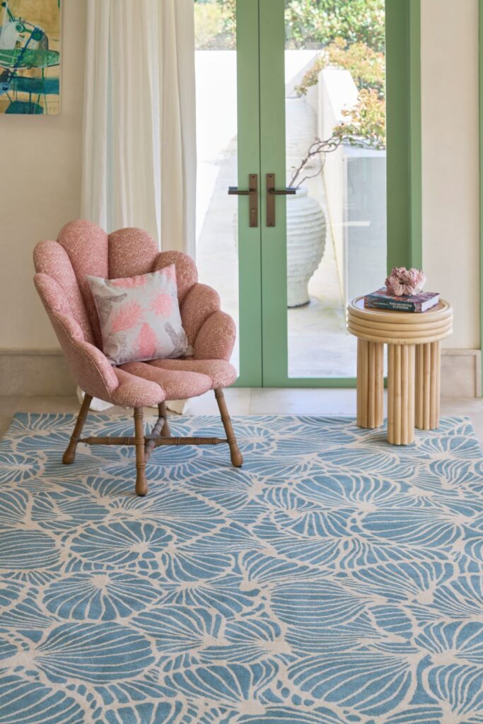 A cozy corner featuring a pink accent chair with floral pillows, a light blue floral rug, and a bamboo-style side table with books, paired with green doors and natural light.