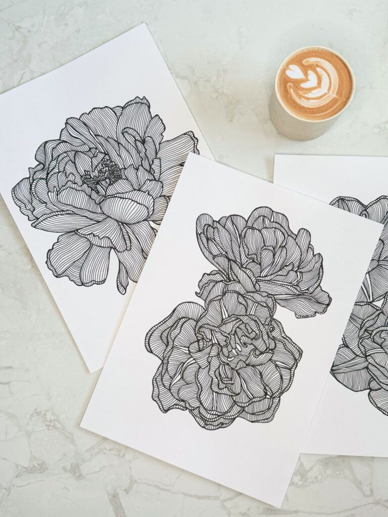 Black and white line art of peony flowers on paper, with a latte featuring a heart design beside them, set on a marble surface.