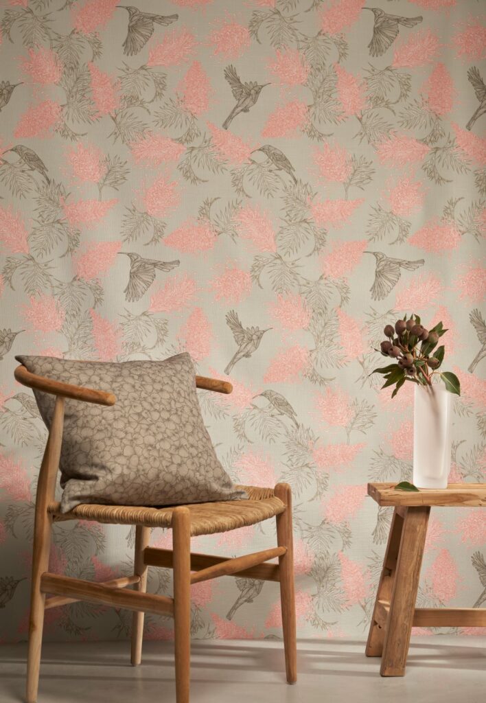 Amalfi-Lines Grevillea_Nectar wallpaper with a soft pink and neutral bird and floral design, paired with a wooden chair, patterned cushion, and a vase with foliage.