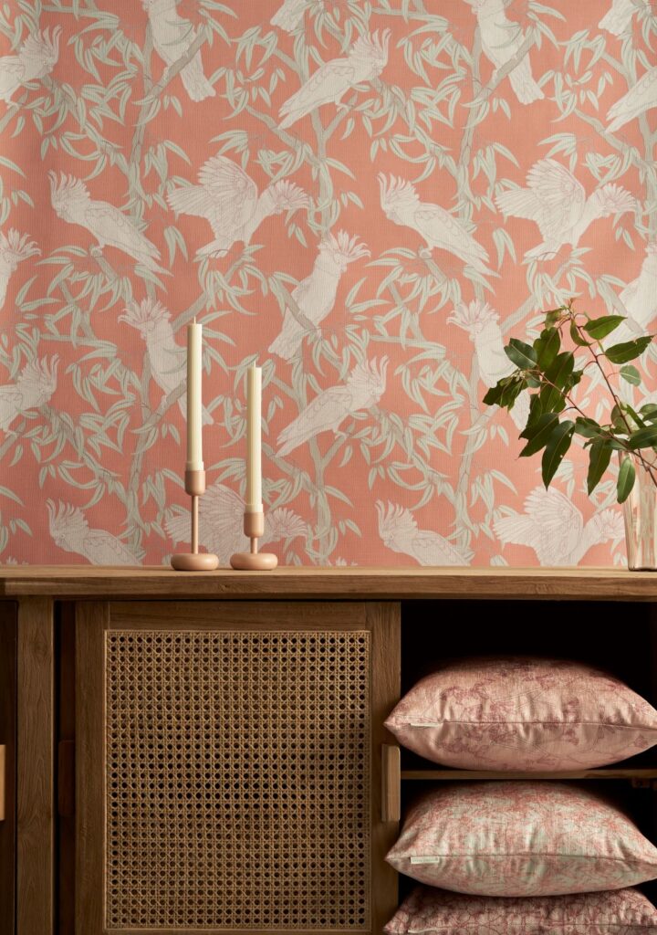 Amalfi-Lines Grevillea_Nectar wallpaper with a soft pink and neutral bird and floral design, paired with a wooden chair, patterned cushion, and a vase with foliage.