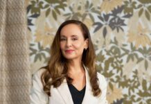 Patricia Braune, a skilled interior designer and surface artist, stands confidently in a white blazer against a botanical print wallpaper, showcasing her design expertise.